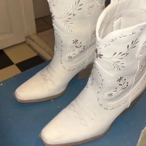 Vita cowboy boots med broderi - Snygga vita cowboy boots med spetsig tå och låg klack. Skorna har broderade blommor och dekorativa sömmar på skaftet. Tillverkade i skinnimitation med träfärgad klack. Perfekta för dig som vill sticka ut med western-vibe.
