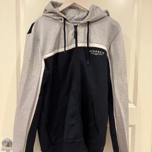 McKenzie hoodie i grått och svart - Snygg hoodie från McKenzie i svart och ljusgrått med vit detalj. Tröjan har huva med snörning, dragkedja framtill och fickor. Perfekt för en chill och sportig stil. Långärmad och i mjukt material som känns skönt mot huden.