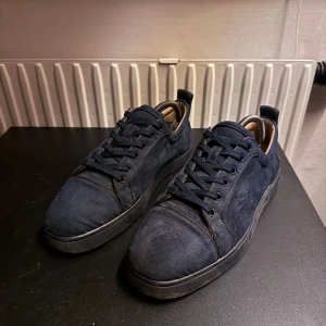 Mörkblå mockasneakers från Louboutin - Snygga mörkblå sneakers i mocka från Christian Louboutin med klassisk röd sula och broderad logga på hälen. Skorna har rund tå, snörning och en stilren design som sticker ut med exklusiva detaljer och lyxig känsla. Passar även 42, har använts en del och har slitage under sulan och i hälen.