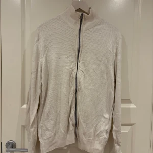 Beige stickad kofta med dragkedja - Säljer en beige stickad kofta med hög krage och hel dragkedja framtill. Tröjan har ett diskret prickigt mönster i stickningen och ribbade muddar vid krage, ärmslut och nederkant. Perfekt för lager-på-lager och chill stil.