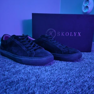 Svarta mockasneakers från Skolyx - Snygga svarta sneakers från Skolyx i mjuk mocka med klassisk rund tå och platt sula. Skorna har snörning och en stilren design som passar perfekt till jeans eller chinos. Levereras med originalkartong.
