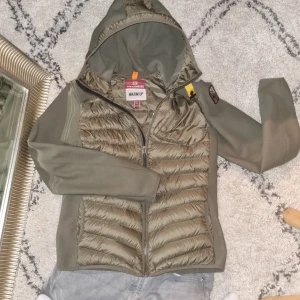 Olivgrön pufferjacka Parajumpers - Snygg olivgrön pufferjacka från Parajumpers med huva och dragkedja. Jackan har quiltade partier framtill och baktill, samt släta ärmar i syntetmaterial. Perfekt för kyliga dagar och har en sportig vibe.