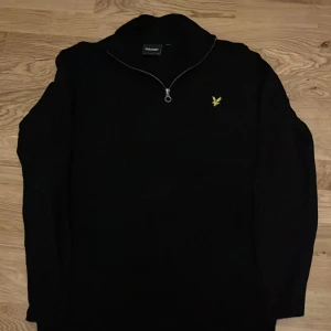 Svart half zip tröja från Lyle & Scott - Svart långärmad tröja från Lyle & Scott med gul broderad logga på bröstet. Tröjan har en snygg half zip-dragkedja vid halsen och är tillverkad i mjuk bomull. Perfekt för dig som gillar stilrena och klassiska plagg med en modern touch.