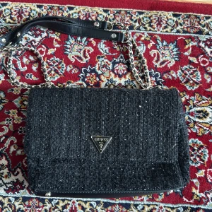 Svart axelväska från Guess - Snygg svart axelväska från Guess med kedjerem och läderdetaljer. Väskan har en mjuk, quiltad yta och ett metalliskt Guess-emblem framtill.  Köpt på zalando för nått år sen för 875kr