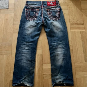 True Religion jeans med röd stitch - Säljer ett par blå jeans från True Religion med tydliga slitningar och coola röda kontrastsömmar. Klassiska bakfickor med märkets signaturbrodyr och patch i rött. Jeansen har rak passform och är tillverkade i denim. Storlek 33 (midja) Skriv gärna om du har frågor eller funderingar!