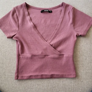 Ljuslila omlottopp från Bik Bok - Superfin ljuslila omlottopp från Bik Bok med korta ärmar och croppad passform. Toppen har en djup v-ringning och är gjord i mjuk bomull.