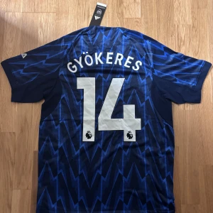 Arsenal Gyökeres #14 Bortatröja Adidas - Snygg Arsenal bortatröja från Adidas med Gyökeres och nummer 14 på ryggen. Tröjan är blå med mörkblå och ljusblå blixtmönster, röda ränder på axlarna och vita tryck. Tillverkad i lätt och ventilerande polyester, perfekt för match eller träning.
