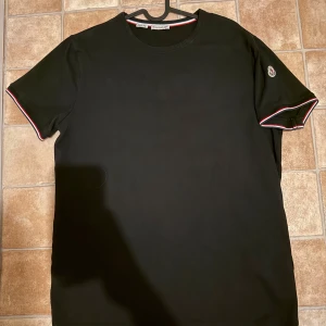 Svart Moncler t-shirt med röd/vit kant - Svart t-shirt från Moncler med korta ärmar och rund hals. Snygga röda och vita detaljer på ärmslut och halsringning. Moncler-logga på vänster ärm ger en exklusiv vibe. Perfekt för dig som gillar stilrena plagg med en twist.