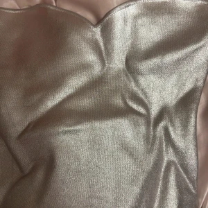 Silver axelbandslös topp från Zara - Snygg axelbandslös topp från Zara i skimrande silverfärgat tyg. Toppen har en hjärtformad överdel och är perfekt för dig som vill sticka ut med en glittrig look. Materialet är mjukt och följsamt, vilket gör den bekväm att bära hela kvällen.