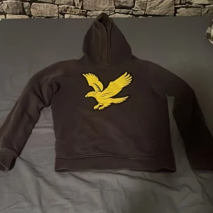 Mörkblå hoodie med gul örn Lyle & Scott - Snygg mörkblå hoodie från Lyle & Scott med stor gul örn på bröstet. Tröjan har huva och långa ärmar, tillverkad i mjuk bomull och polyesterblandning. Perfekt för dig som gillar streetwear och vill sticka ut med en cool logga.