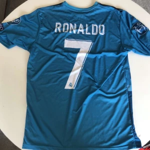 Real Madrid Ronaldo #7 blå fotbollströja - Säljer en blå Real Madrid fotbollströja,  Adidas med Ronaldo och nummer 7 på ryggen. Tröjan har korta ärmar, CL-märke på ärmen, Fly Emirates-tryck och pixelmönster framtill. Materialet är lätt och andas, perfekt för match, träning eller vardagligt bruk.