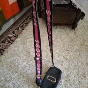 Marc Jacobs svart axelväska med bred rem - Cool svart axelväska från Marc Jacobs med bred, justerbar rem i rosa och svart med logga. Väskan har guldiga detaljer, dubbla dragkedjor och röd insida. Perfekt storlek för det viktigaste och ger en trendig vibe till din outfit.