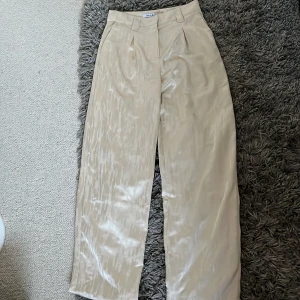 Beiga vida byxor i satin från NA-KD - Snygga beiga byxor från NA-KD i glansigt satinmaterial. Vida ben och hög midja ger en trendig siluett. Byxorna har pressveck och bälteshällor för extra stilpoäng. Perfekta för dig som gillar en chic och modern look.