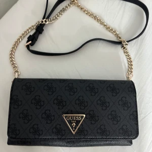 Svart axelväska från Guess med kedja - Snygg svart axelväska från Guess med silver kedja och läderrem. Väskan har Guess-logga framtill, diskret mönster, dragkedja på baksidan och flera fack för kort och småsaker inuti. Perfekt storlek för det viktigaste och riktigt stilren look. Väskan har aldrig använt. 