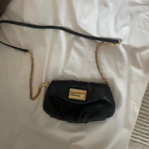 Svart axelväska Marc by Marc Jacobs - Snygg svart axelväska från Marc by Marc Jacobs i mjukt skinn med guldfärgade kedjedetaljer och lång axelrem. Väskan har ett coolt innerfoder med stora bokstäver och en guldig logoplatta framtill. Perfekt storlek för det viktigaste.