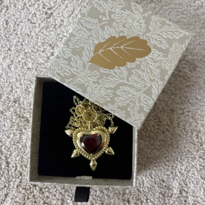Guldigt halsband med hjärthänge - Unikt Ioaku halsband i guldton med ett stort hjärthänge i röd sten och coola spetsiga detaljer runt om. Kedjan är tunn och har en liten bricka med läppmotiv vid låset. Perfekt statement-piece för dig som vill sticka ut. Köpt för 900kr säljer för 350kr, aldrig använt. Pris kan diskuteras. 