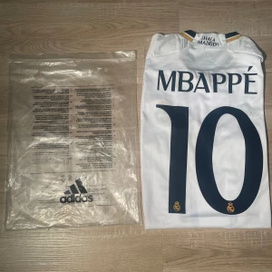 Real Madrid Mbappé #10 Adidas tröja - Säljer en officiell Real Madrid fotbollströja från Adidas med Mbappé och nummer 10 på ryggen. Tröjan är vit med mörkblå detaljer, guldiga ränder på axlarna och Champions League-märke på ärmen. Materialet är lätt och andas, perfekt för match eller träning.