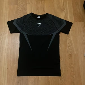 Svart Gymshark t-shirt med mönster - Snygg svart t-shirt från Gymshark i storlek S. Tröjan har korta ärmar, grått grafiskt mönster och diskret logga på bröstet. Tillverkad i ett stretchigt syntetmaterial som andas, perfekt för träning och aktiva dagar.