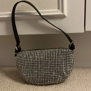 Glittrig axelväska med strass - Säljer en superglittrig axelväska täckt av silvriga strasstenar. Väskan har svart bas, rundad form och ett svart axelband. Stängs med dragkedja upptill. Perfekt för dig som vill sticka ut och addera extra bling till din outfit. Samtliga stenar sitter kvar!