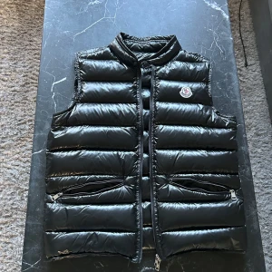 Moncler väst - Snygg svart dunväst från Moncler med glansig finish och klassisk logga på bröstet. Västen har ståkrage, två dragkedjefickor och quiltad design. Perfekt att slänga över en hoodie eller tröja för extra värme och stilpoäng. Äktehetsbevis på bild 4 och 5. Monclers storlek 1 sitter som xs/s.