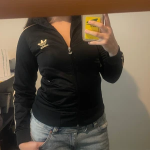 Svart Adidas hoodie med guldiga ränder - Svart hoodie från Adidas med klassiska guldiga tre ränder längs ärmarna och Adidas-logga på bröstet. Tröjan har dragkedja framtill, huva och en adidas logga på ryggen. 