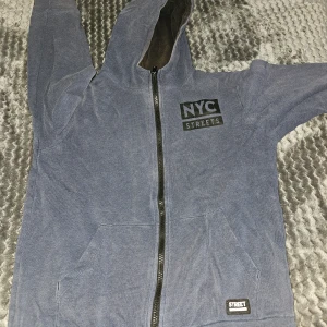 Blå NYC hoodie med dragkedja - Säljer en blå hoodie från NYC Streets med tryck både fram och bak. Tröjan har huva, lång ärm och praktisk dragkedja hela vägen. Mjuk bomullskänsla och fickor framtill. Perfekt för dig som gillar streetstyle och vill ha en chill look.