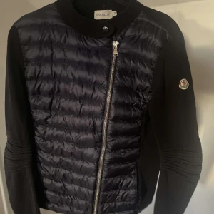 Mörkblå cardigan jacka från Moncler - Moncler Cardigan Jacket – Dam strl M –  mörkblå quiltad front & stickade ärmar Elegant och mångsidig Moncler cardigan/jacket i mörkblå, perfekt för övergångssäsonger eller som ett lättare ytterplagg.🎨 Färg: mörkblå 📏 Storlek: M (regular fit, passar ca EU 38–40) 📐 Mått: Längd ca 58 cm, ärmlängd ca 63 cm, bröstvidd ca 48 cm (platt mått) ♻️ Skick: Mycket bra begagnat skick – välvårdad med minimala spår av användning 🧵 Material: Nylon med lätt dunfyllning samt stickat ullblanding ärmar 