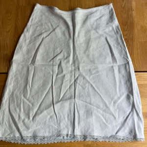 Vit långkjol med spetskant - Säljer en enkel och stilren vit långkjol med mjukt tyg och snygg spetsdetalj längst ner. Kjolen har rak passform och är perfekt för dig som gillar minimalistisk stil med en touch av romantik. Spetsen ger kjolen en extra charmig vibe. Materialet är förmodligen linne men finns ej någon lapp.