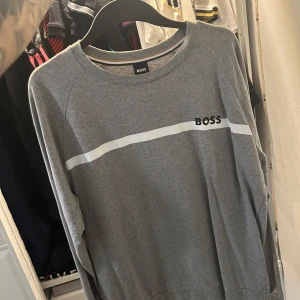 Grå långärmad tröja från BOSS - Säljer en stilren grå långärmad tröja från BOSS med vit horisontell rand och logga på bröstet. Tröjan har rund halsringning och är tillverkad i mjuk bomull, perfekt för en clean och avslappnad look.