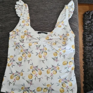 Vit blommig linnetopp från Mohito - Superfin vit topp från Mohito med citrongult blommönster och volangprydda axelband. Toppen har en söt twistad detalj framtill och är perfekt för varma dagar. Luftigt material och romantisk vibe.