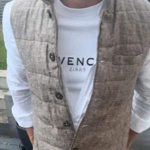 John Henric väst - Säljer en beige quiltad dunväst med snygg linnestruktur och knäppning framtill. Västen har en stilren look med diskreta sömmar och en liten bröstficka. Perfekt att slänga över en skjorta för en clean och modern vibe.