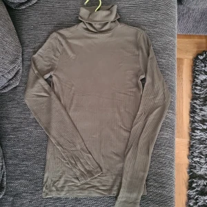 Ribbad olivgrön polotröja från New Yorker - Snygg ribbad polotröja från New Yorker i en olivgrön nyans. Tröjan har lång ärm och hög polokrage, perfekt för lager-på-lager. Materialet är mjukt och stretchigt, vilket gör den skön att bära. Passar dig som gillar stilrena och enkla plagg.