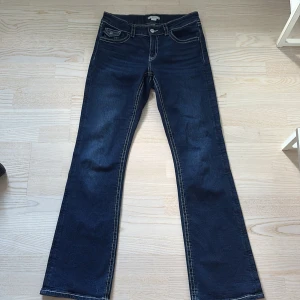 Mörkblå bootcut jeans med kontrastsömmar - Snygga mörkblå jeans med bootcut-snitt och tydliga vita kontrastsömmar. Byxorna har klassiska fickor fram och bak, där bakfickorna har lock och knappdetalj. Perfekta för dig som gillar en tidlös och cool look.