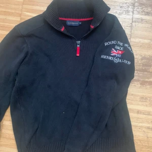 Svart stickad tröja Henri Lloyd half zip - Svart stickad tröja från Henri Lloyd med broderad logga och brittisk flagga på ärmen. Tröjan har en hög krage med röda detaljer och half zip-dragkedja. Perfekt för dig som gillar klassisk och sportig stil.