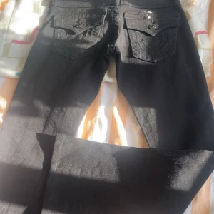 Svarta jeans från True Religion, stl 24 - Säljer ett par svarta jeans från True Religion med snygga detaljerade bakfickor och klassiska knappar. Jeansen har en rak passform och är tillverkade i jeansmaterial. Perfekta för dig som gillar en stilren look med lite edge.