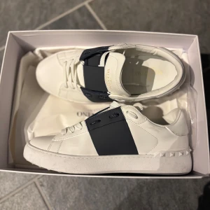 Valentino Garavani Open Sneakers vit/blå - Snygga Valentino Garavani Open Sneakers i vitt kalvskinn med bred mörkblå rem över vristen och ikoniska nitar på hälen. Klassisk rund tå, platt sula och vita snören. Perfekta för dig som vill ha en clean men lyxig look.