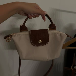 Beige och brun handväska Longchamp - Snygg beige handväska från Longchamp med bruna skinnhandtag och detaljer. Väskan har guldiga metalldetaljer, dragkedja och ett klassiskt lock med tryckknapp.