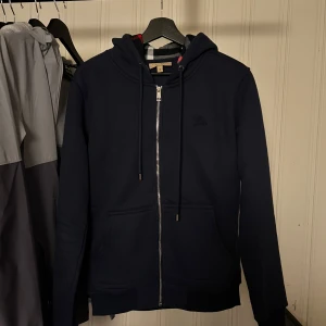 Mörkblå hoodie från Burberry med dragkedja - Säljer en mörkblå hoodie från Burberry i storlek M. Använd fåral gånger. 10/10 Skick! Tröjan har hel dragkedja, huva med snörning och klassiskt rutigt foder i luvan. Tillverkad i mjuk bomull med lite elastan för extra komfort. Perfekt för en stilren och avslappnad look.