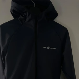 Svart vindjacka från Sail Racing - Säljer en svart vindjacka från Sail Racing med huva och dragkedja framtill. Jackan har en stilren look med logga på bröstet och är tillverkad i ett slitstarkt syntetmaterial. Perfekt för blåsiga dagar och enkel att matcha med det mesta.