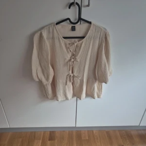 Beige blus med knyt fram från H&M - Superfin beige blus med puffiga halvlånga ärmar och tre knyt framtill. Tillverkad i mjuk bomull med lätt skrynklig struktur. Perfekt för dig som gillar romantiska detaljer och avslappnad stil.
