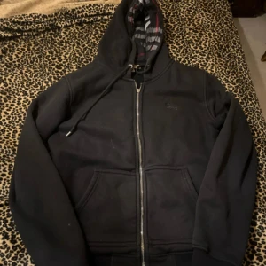 Svart hoodie från Burberry med dragkedja - Svart hoodie från Burberry i storlek M med klassisk dragkedja framtill och snörning i huvan. Insidan av huvan har Burberrys rutiga mönster i svart, grått och rött. Broderad logga på bröstet och två stora fickor framtill. Perfekt för en chill och stilren look.