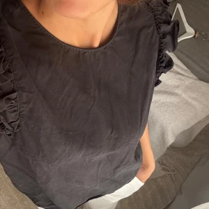 Svart blus med volangärmar och knyt - Säljer en svart blus med volangdetaljer på ärmarna och  knytning i nacken som gör halsen lite bredare och djupare där bak. Blusen är kortärmad och har en rak passform. Perfekt för dig som gillar stilrena plagg med en twist. Materialet känns mjukt och lätt, troligen bomull.