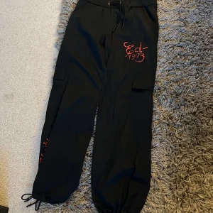 Svarta cargopants från Ed Hardy med drake - Svarta cargopants från Ed Hardy med röd logga och broderad drake på benet. Byxorna har snörning i midjan och vid benslut, samt stora fickor på sidorna. Knappt använda!
