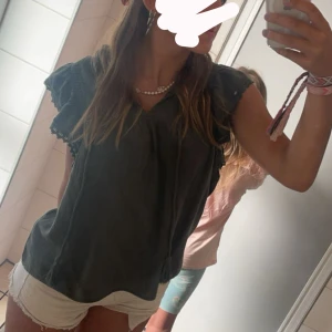 Mörkgrå blus med volang från Pigalle - Supersöt mörkgrön blus från Pigalle med volangdetaljer på ärmarna och knytband vid halsen. Luftig och skön modell i mjukt tyg, perfekt till shorts eller jeans. Ärmlös design med snygga virkade detaljer på axlarna.