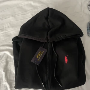Svart hoodie från Polo Ralph Lauren - Svart hoodie från Polo Ralph Lauren med dragkedja och huva. Klassisk röd logga broderad på bröstet, justerbara snören i huvan och känguruficka framtill. Perfekt för en chill och stilren look.