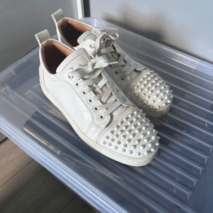 Vita Christian Louboutin sneakers med nitar - Säljer ett par vita sneakers från Christian Louboutin med coola nitar över tån och klassisk röd sula. Skorna har snörning och är gjorda i skinn med en rund tå. Perfekt för dig som vill sticka ut med en edgy vibe och exklusiv känsla. Dem e mycket bra skick förutom att dem e smutsiga