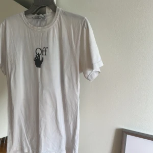 Vit Off-White t-shirt med tryck - Säljer en clean vit t-shirt från Off-White i bomull, med svart hand- och texttryck på bröstet och stor grafisk logga i blått och gult på ryggen. Klassisk rund hals och korta ärmar. Perfekt för dig som gillar streetwear och vill sticka ut med snygga detaljer.