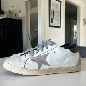 Golden Goose Superstar sneakers vit/svart - Golden Goose Superstar sneakers i vitt skinn med grå mockadetaljer och svart häl. Grå snörning och ikonisk stjärna på sidan. Platt sula och rund tå. Snyggt sliten look och cool design som sticker ut. Perfekt för dig som gillar streetstyle och italiensk mode. Säljer pga för små.