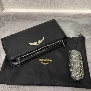 Svart axelväska från Zadig & Voltaire - Snygg svart axelväska från Zadig & Voltaire i skinn med silverfärgad vinge framtill. Väskan har en stilren design med dragkedja och medföljande axelrem i kedja. Perfekt för dig som gillar en edgy och modern look.