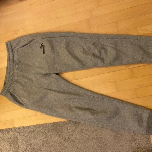 Grå mjukisbyxor från Puma - Säljer ett par grå mjukisbyxor från Puma med elastisk midja och muddar vid bensluten. Byxorna har en klassisk logga på vänster lår och är tillverkade i mjukt material som är perfekt för chill eller träning. Snygg och enkel design med fickor på sidorna. Pris kan diskuteras!